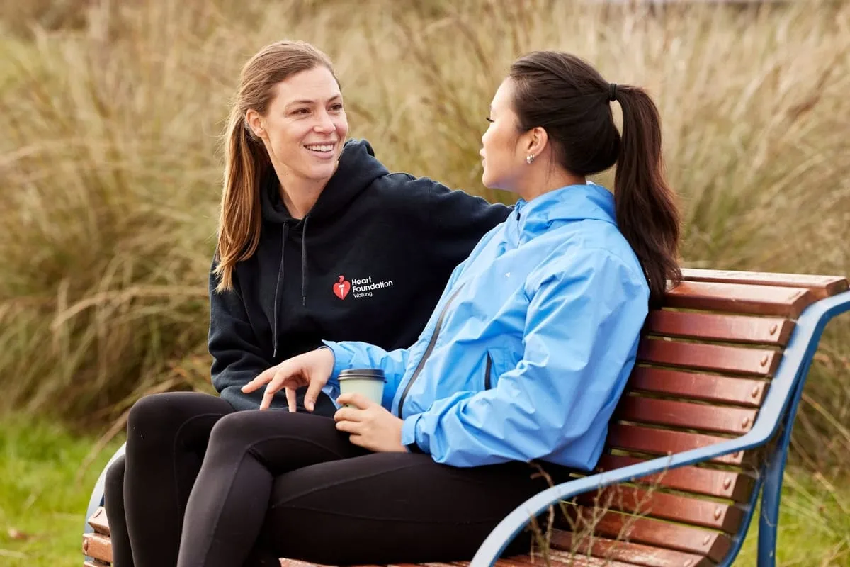 Two women talking on a park bench, one in a Heart Foundation Walking hoodie.